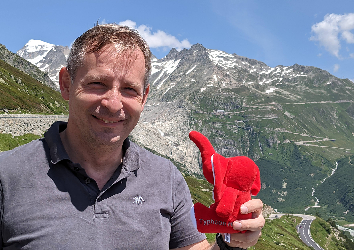 A man standing in front of mountains, holding a stuffed elephant and smiling.