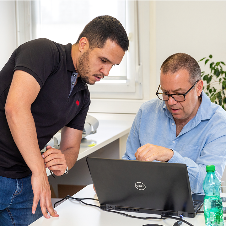 One man focused on a laptop screen, and another standing besides and helping him.